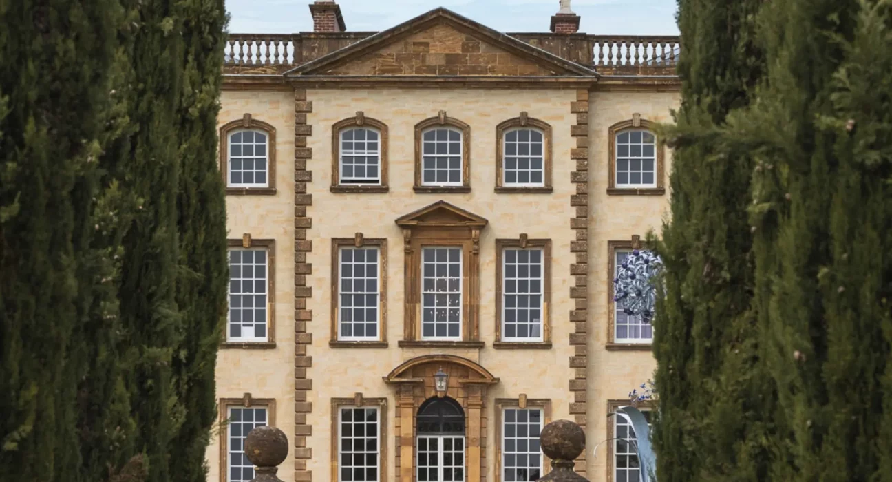 Sherborne House, a Grade I listed Georgian mansion in golden stone, framed by tall cypress trees, with sculptural artwork visible in one of its windows