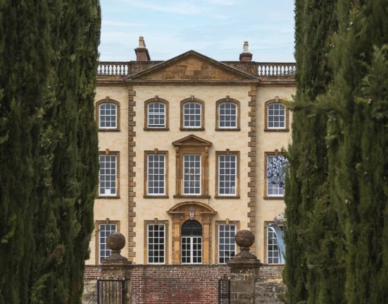 Sherborne House, a Grade I listed Georgian mansion in golden stone, framed by tall cypress trees, with sculptural artwork visible in one of its windows
