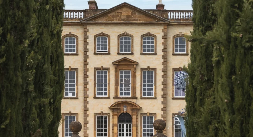 Sherborne House, a Grade I listed Georgian mansion in golden stone, framed by tall cypress trees, with sculptural artwork visible in one of its windows