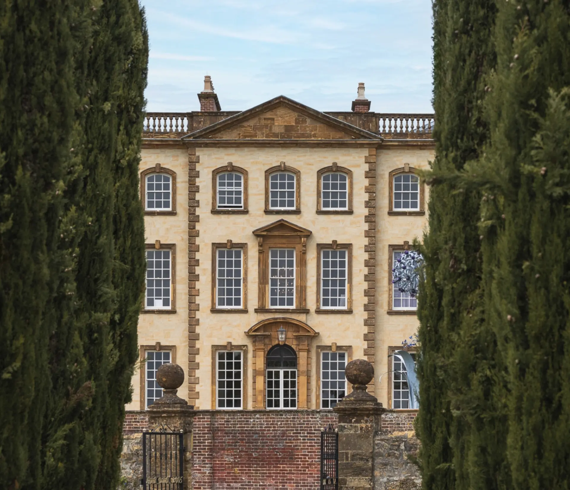 Sherborne House, a Grade I listed Georgian mansion in golden stone, framed by tall cypress trees, with sculptural artwork visible in one of its windows