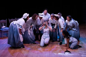 Women of Dorset performers in period costume gather around a kneeling figure holding an object to her face, on a stage scattered with flour