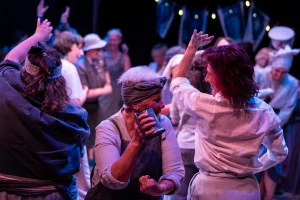 Women of Dorset performers in period costume dance and celebrate together on stage under blue and purple lighting, with one holding a tankard