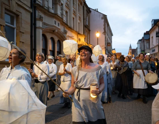 "Women of Dorset lantern parade through Dorchester streets at dusk, carrying handmade paper lanterns and candles in vintage-style dress, singing together