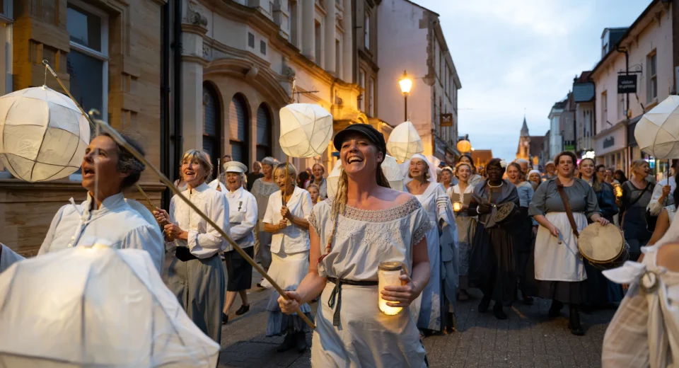"Women of Dorset lantern parade through Dorchester streets at dusk, carrying handmade paper lanterns and candles in vintage-style dress, singing together