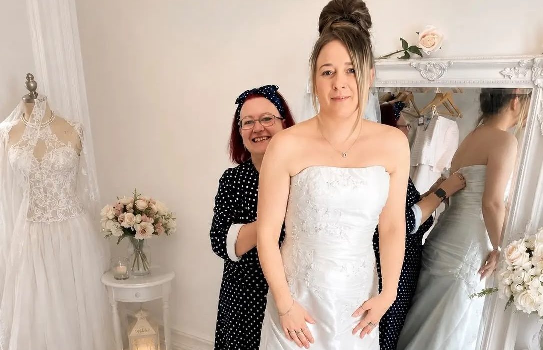 Alt text: "Bridal Floor volunteer Nicky Cullis fits a bride into a strapless white wedding dress on a fitting platform at Scope's Bridport shop, with shop manager Sonia Hallett's reflection visible in a large mirror beside racks of dresses