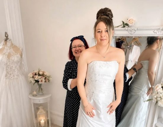 Alt text: "Bridal Floor volunteer Nicky Cullis fits a bride into a strapless white wedding dress on a fitting platform at Scope's Bridport shop, with shop manager Sonia Hallett's reflection visible in a large mirror beside racks of dresses