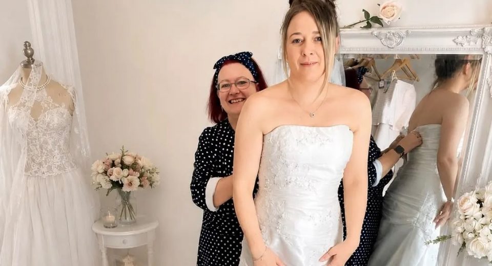 Alt text: "Bridal Floor volunteer Nicky Cullis fits a bride into a strapless white wedding dress on a fitting platform at Scope's Bridport shop, with shop manager Sonia Hallett's reflection visible in a large mirror beside racks of dresses