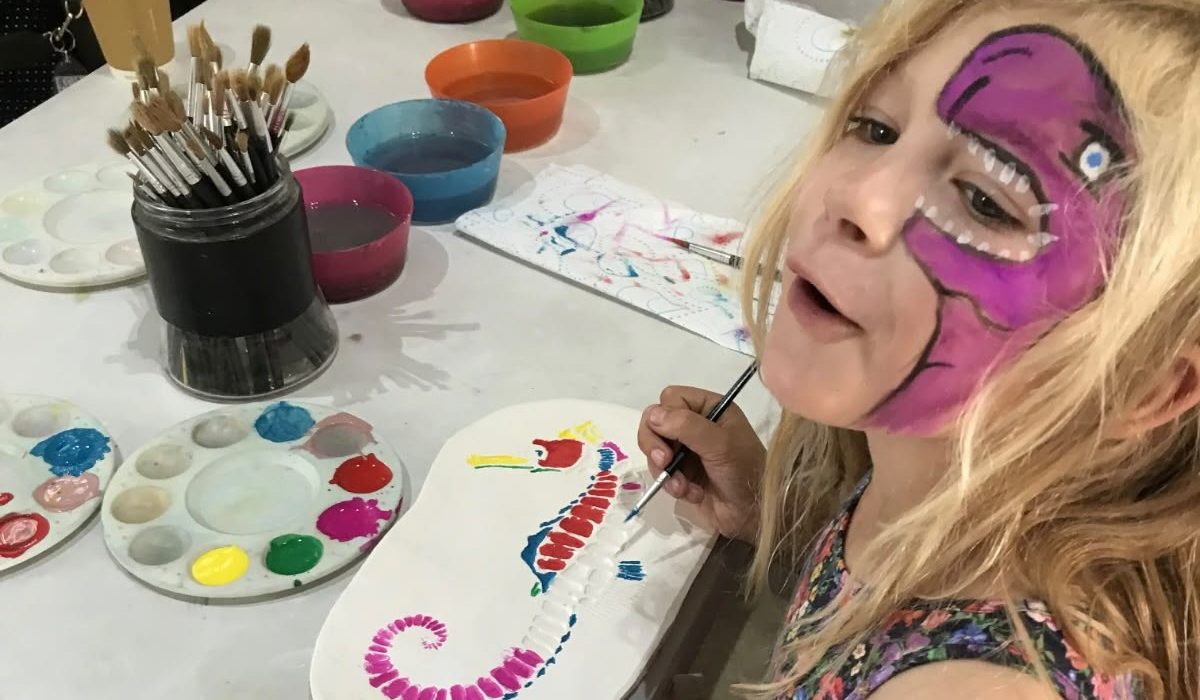 A young girl with a purple dinosaur face paint design smiles at the camera while painting a colourful seahorse fossil ornament at a craft table during a family event at Portland Museum. The table is covered with paint palettes, brushes, and bowls of paint water in red, green, blue and orange