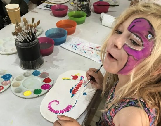 A young girl with a purple dinosaur face paint design smiles at the camera while painting a colourful seahorse fossil ornament at a craft table during a family event at Portland Museum. The table is covered with paint palettes, brushes, and bowls of paint water in red, green, blue and orange