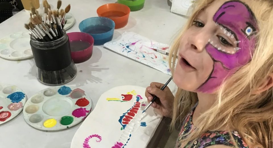 A young girl with a purple dinosaur face paint design smiles at the camera while painting a colourful seahorse fossil ornament at a craft table during a family event at Portland Museum. The table is covered with paint palettes, brushes, and bowls of paint water in red, green, blue and orange