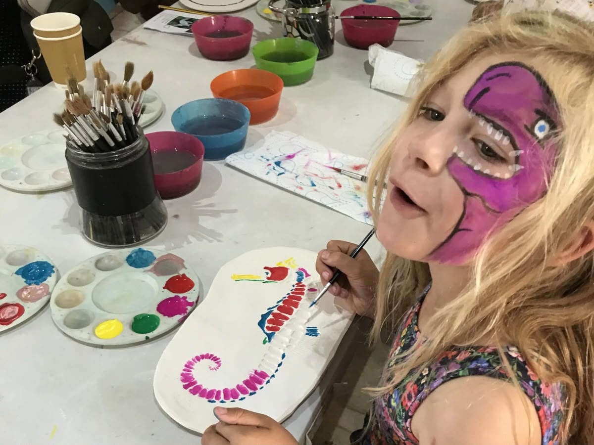 A young girl with a purple dinosaur face paint design smiles at the camera while painting a colourful seahorse fossil ornament at a craft table during a family event at Portland Museum. The table is covered with paint palettes, brushes, and bowls of paint water in red, green, blue and orange