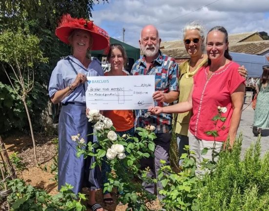 Five smiling people stand among white rose bushes at Bridport Vintage Market, holding a large Barclays cheque for £1,000 made out to Bridport Food Matters.