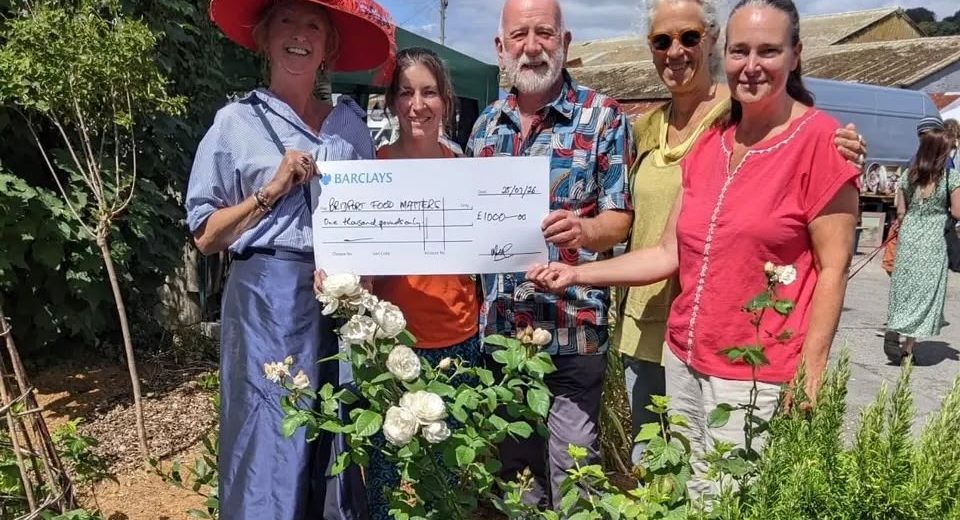Five smiling people stand among white rose bushes at Bridport Vintage Market, holding a large Barclays cheque for £1,000 made out to Bridport Food Matters.