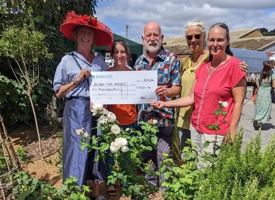 Five smiling people stand among white rose bushes at Bridport Vintage Market, holding a large Barclays cheque for £1,000 made out to Bridport Food Matters.