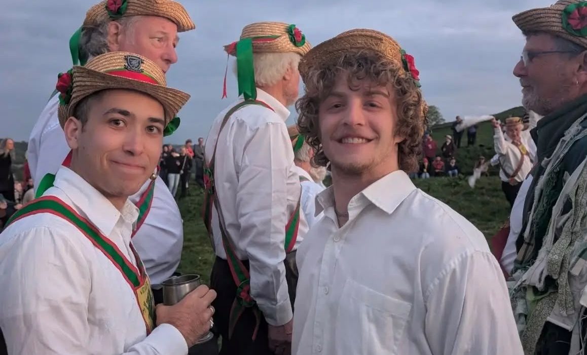 Wessex Morris Men in traditional straw boater hats with ribbons and rosettes gather outdoors on a hillside at dusk, smiling for the camera in white shirts and red-and-green sashes.