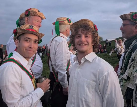 Wessex Morris Men in traditional straw boater hats with ribbons and rosettes gather outdoors on a hillside at dusk, smiling for the camera in white shirts and red-and-green sashes.