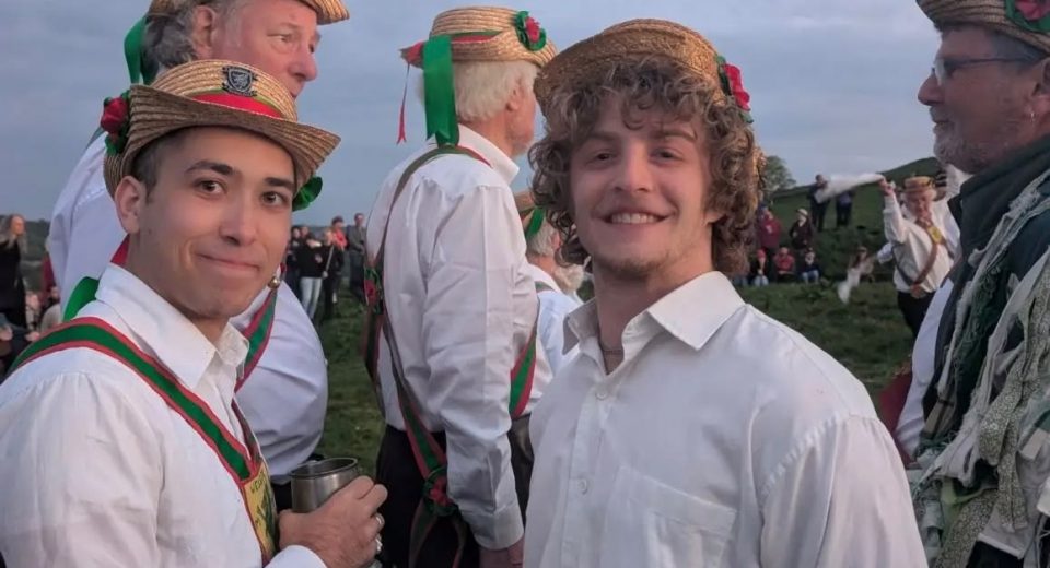 Wessex Morris Men in traditional straw boater hats with ribbons and rosettes gather outdoors on a hillside at dusk, smiling for the camera in white shirts and red-and-green sashes.