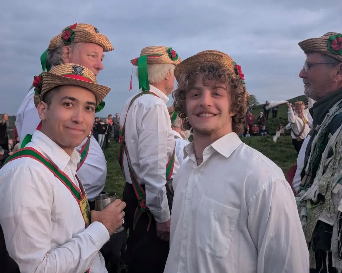 Wessex Morris Men in traditional straw boater hats with ribbons and rosettes gather outdoors on a hillside at dusk, smiling for the camera in white shirts and red-and-green sashes.
