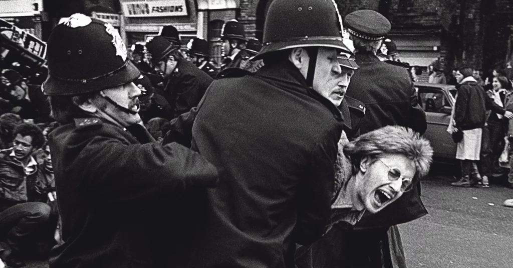 Black and white photojournalism image by Carlos Guarita showing two British police officers in traditional custodian helmets forcibly restraining a woman with curly hair and glasses, her face contorted in a scream, during an Irish demonstration in Kilburn, London. Crowds of onlookers and other officers are visible in the background
