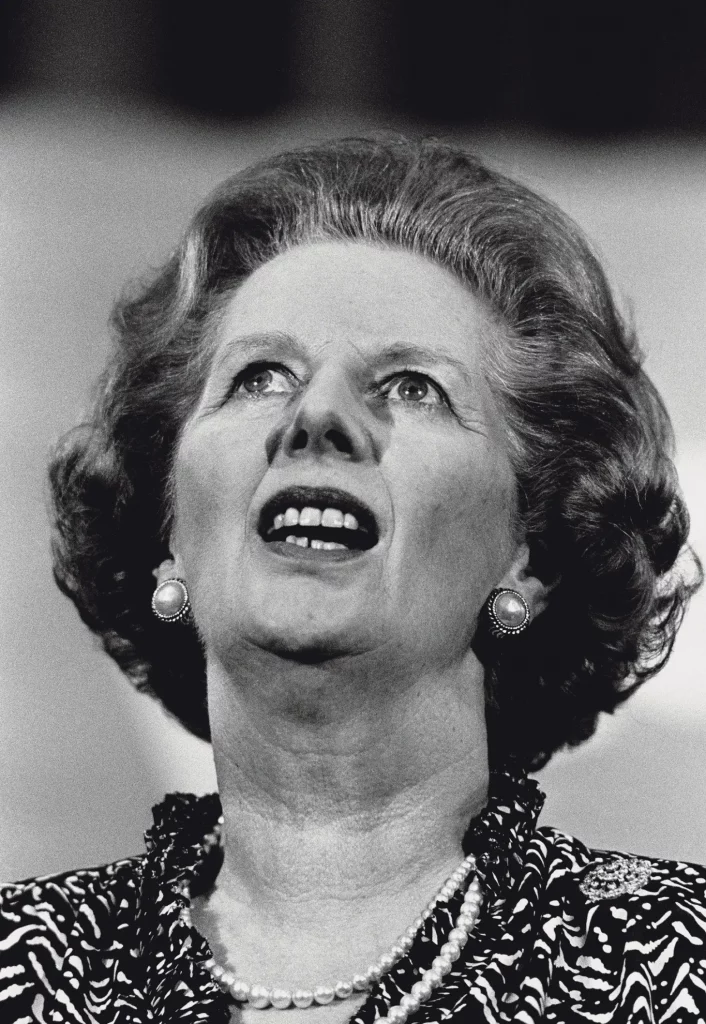 Black and white photojournalism close-up by Carlos Guarita of Margaret Thatcher, wearing pearl earrings and a pearl necklace, looking upward with mouth open mid-speech at the Conservative Women's Conference, Central Hall, Westminster.