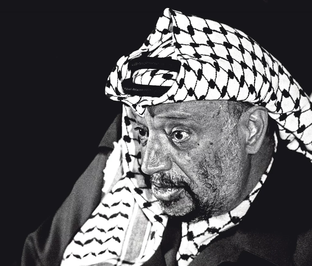Black and white photojournalism close-up by Carlos Guarita of Yasser Arafat wearing a checkered keffiyeh headdress, looking downward with a pensive expression