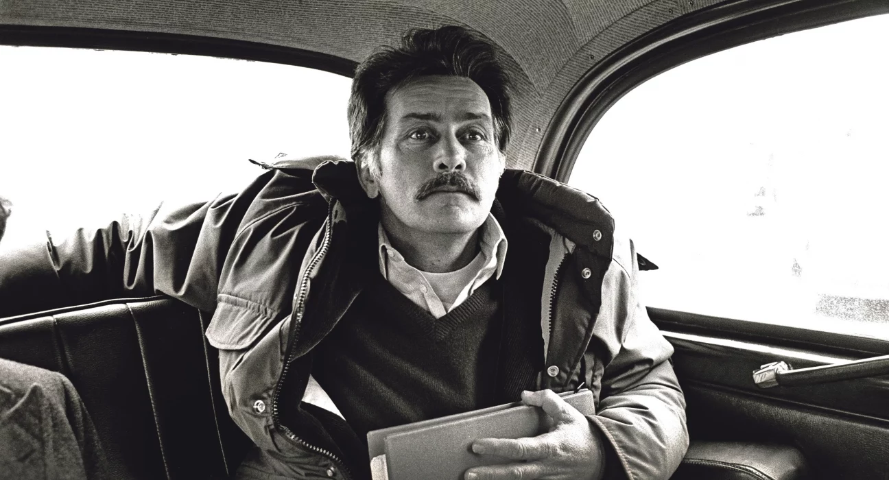 Black and white photojournalism image by Carlos Guarita of actor Martin Sheen sitting in the back of a London taxi, wearing a padded jacket over a jumper and shirt, holding a folder, with a moustache and a thoughtful, slightly averted gaze