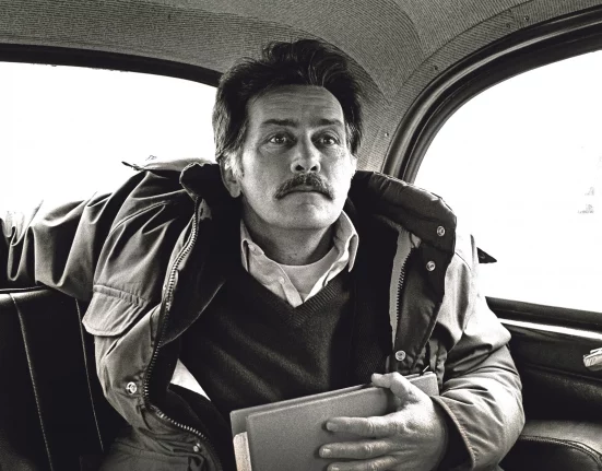 Black and white photojournalism image by Carlos Guarita of actor Martin Sheen sitting in the back of a London taxi, wearing a padded jacket over a jumper and shirt, holding a folder, with a moustache and a thoughtful, slightly averted gaze
