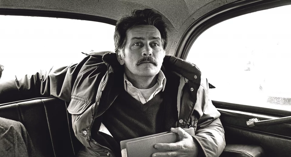 Black and white photojournalism image by Carlos Guarita of actor Martin Sheen sitting in the back of a London taxi, wearing a padded jacket over a jumper and shirt, holding a folder, with a moustache and a thoughtful, slightly averted gaze