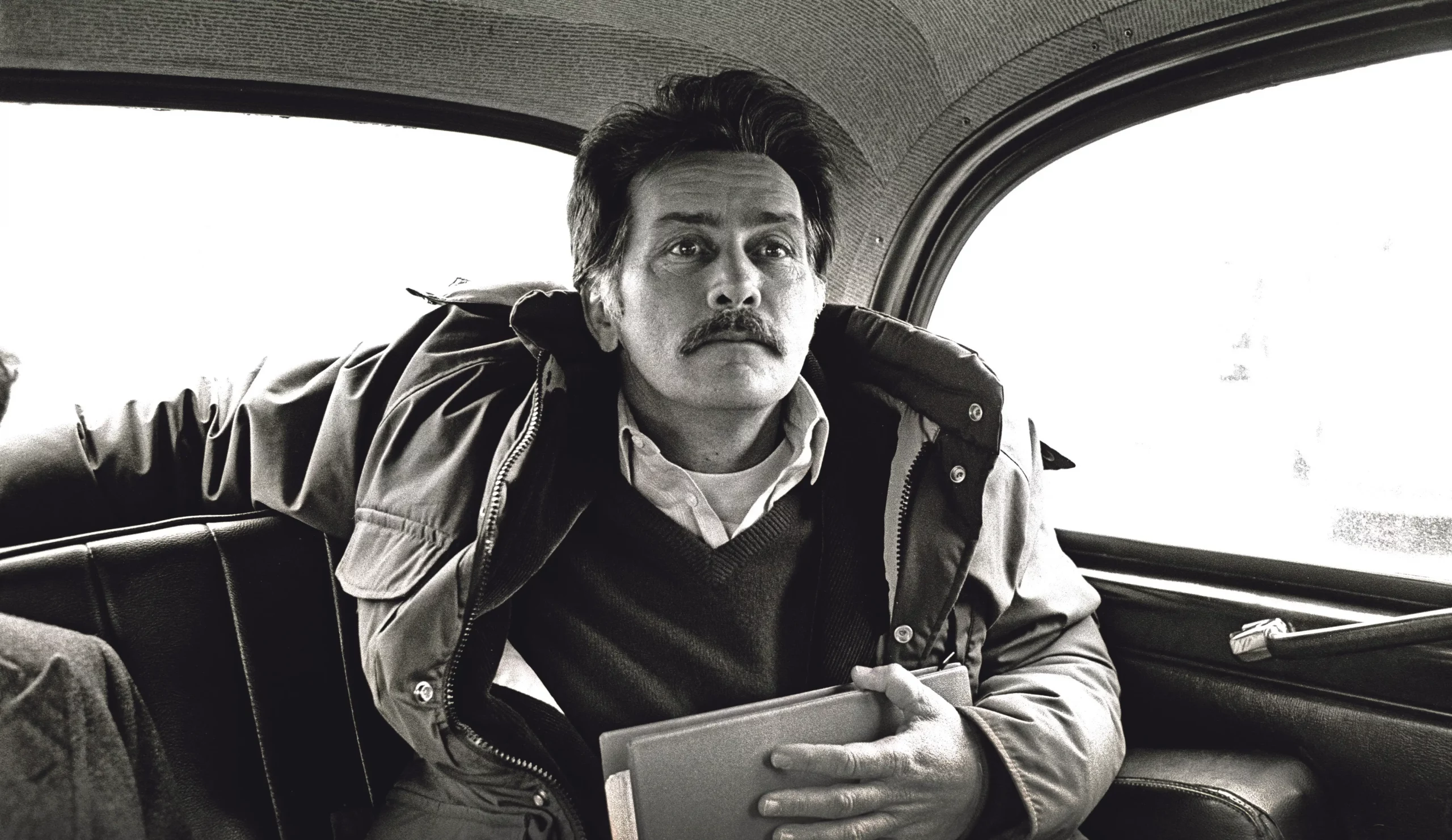 Black and white photojournalism image by Carlos Guarita of actor Martin Sheen sitting in the back of a London taxi, wearing a padded jacket over a jumper and shirt, holding a folder, with a moustache and a thoughtful, slightly averted gaze