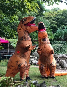 Two people in large inflatable orange T-Rex costumes stand facing each other on the lawn at Portland Museum, roaming the grounds among trees and rockery as part of the Dino Day event