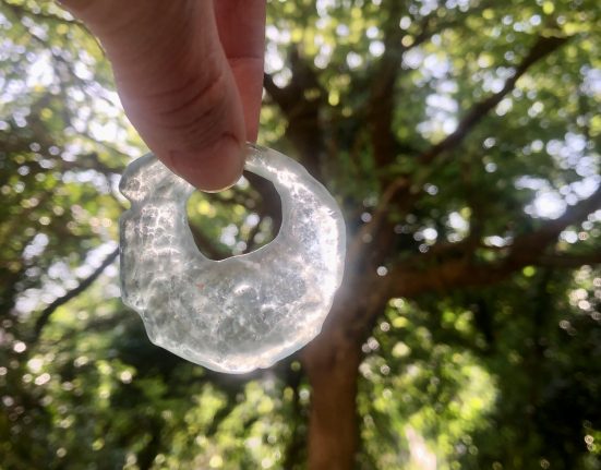 A hand holds up an irregular glass torus pendant made from broken glass found at France Quarry, backlit by sunlight through the woodland canopy