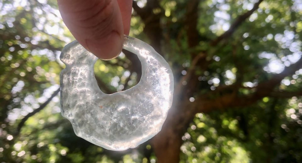 A hand holds up an irregular glass torus pendant made from broken glass found at France Quarry, backlit by sunlight through the woodland canopy