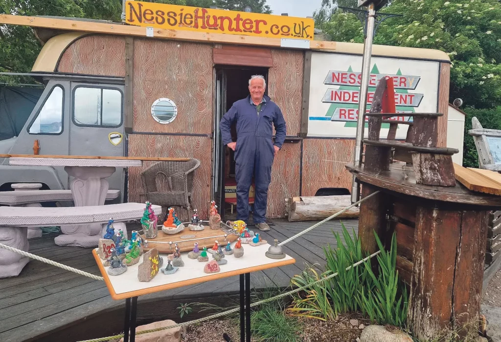 Steve Feltham outside his Nessie Hunter caravan at Loch Ness, with his handmade Loch Ness Monster models displayed on a table