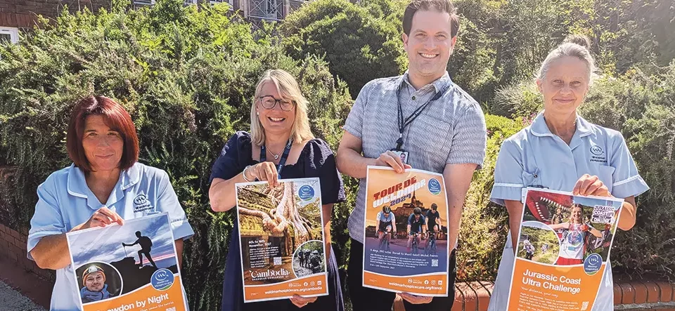 Weldmar Hospicecare staff hold posters for the four new 2027 fundraising challenges - Snowdon by Night, Cambodia Overseas Challenge, Tour de Weldmar and Jurassic Coast Ultra Challenge - outside the hospice in Dorchester