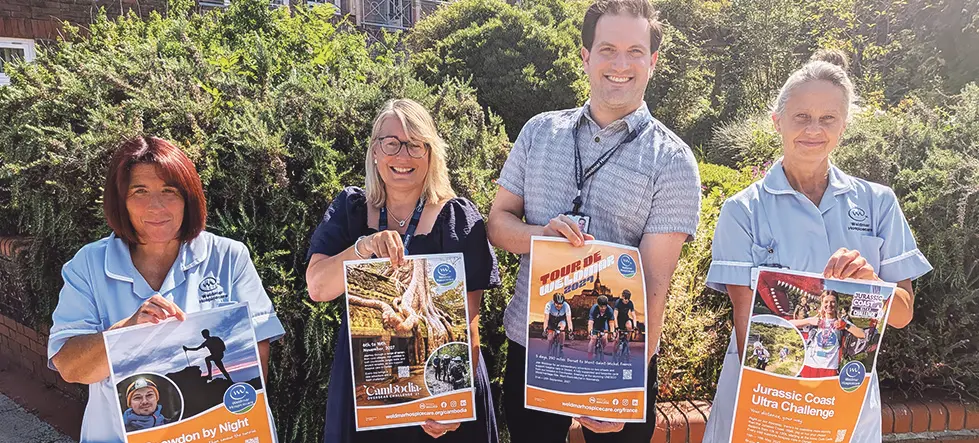 Weldmar Hospicecare staff hold posters for the four new 2027 fundraising challenges - Snowdon by Night, Cambodia Overseas Challenge, Tour de Weldmar and Jurassic Coast Ultra Challenge - outside the hospice in Dorchester