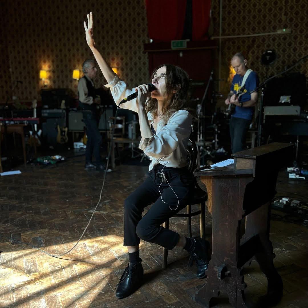 PJ Harvey, author of Orlam, sings into a microphone with one arm raised, seated beside a wooden lectern in a sunlit hall