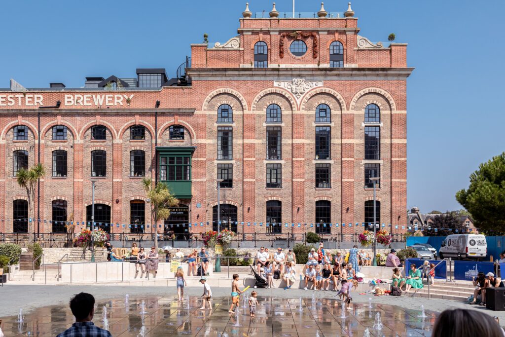 Historic red-brick former Dorchester Brewery building at Brewery Square, with children playing in the splash fountains and visitors relaxing on the terraces below.