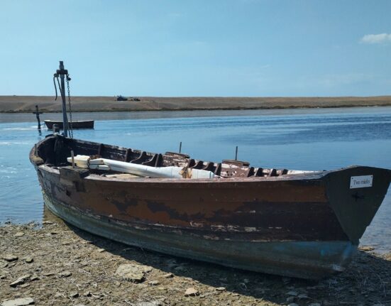 A weathered wooden Fleet trow on the shore of the Fleet lagoon, with Chesil Beach in the background.
