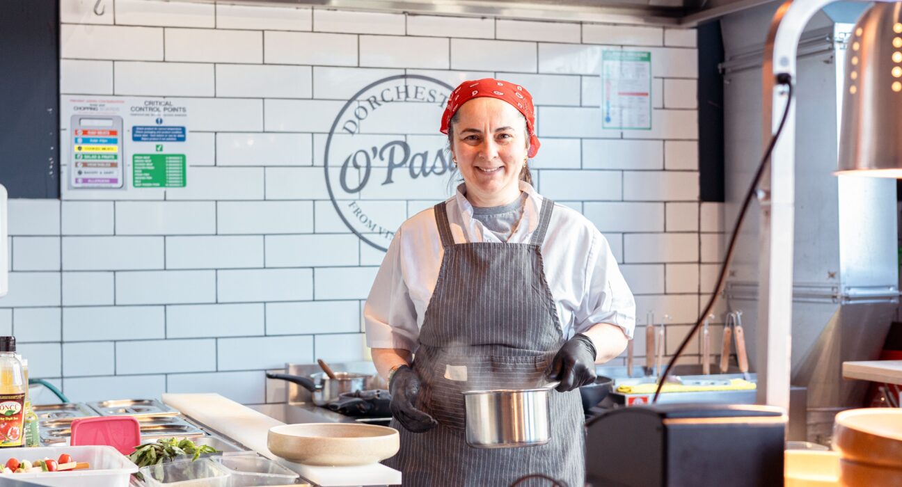 Chef in a red bandana and striped apron smiles while holding a metal pot in the kitchen at O’Pasta, Brewery Square, Dorchester.