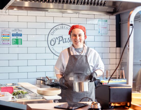 Chef in a red bandana and striped apron smiles while holding a metal pot in the kitchen at O’Pasta, Brewery Square, Dorchester.