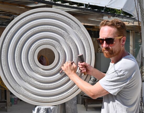 Stone carver Ben Russell uses a mallet and chisel on a large circular limestone sculpture of textured concentric rings in his outdoor workshop.