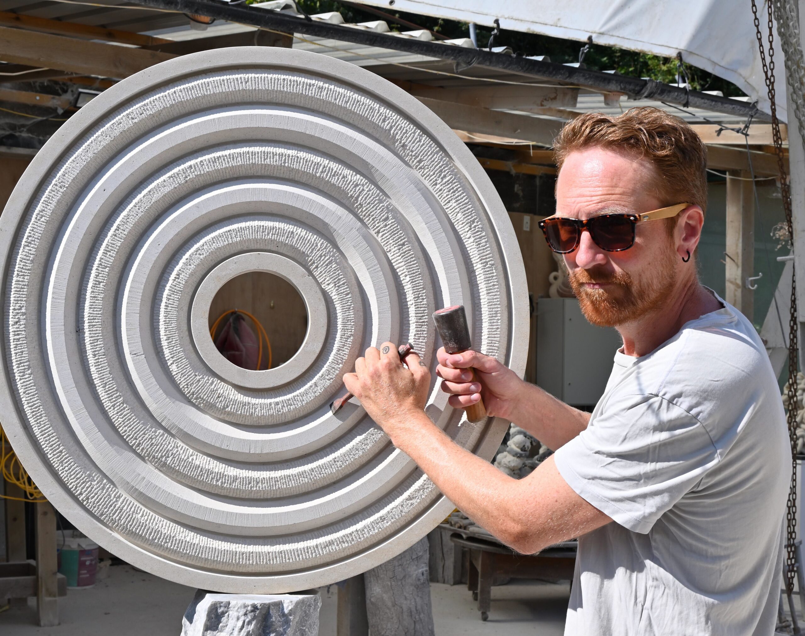 Stone carver Ben Russell uses a mallet and chisel on a large circular limestone sculpture of textured concentric rings in his outdoor workshop.