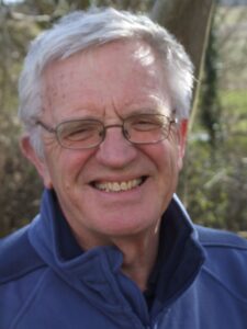 Bob Holman, 82, of West Dorset Magazine, working past retirement age, smiles outdoors in a blue fleece and glasses.