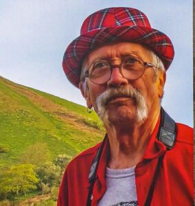 Doug Chalk, 81, photographer behind the Colmer’s Hill calendar, working past retirement age, in a tartan hat with a camera strap against a green hillside.
