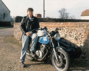 Young Chris Jury, Lovejoy actor, wearing a leather jacket and jeans and sitting on a blue motorbike in front of a flint and brick wall in a village lane.