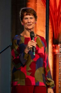 Lois Pearson, 75, director of Beaminster Festival, working past retirement age, speaks into a microphone on stage.