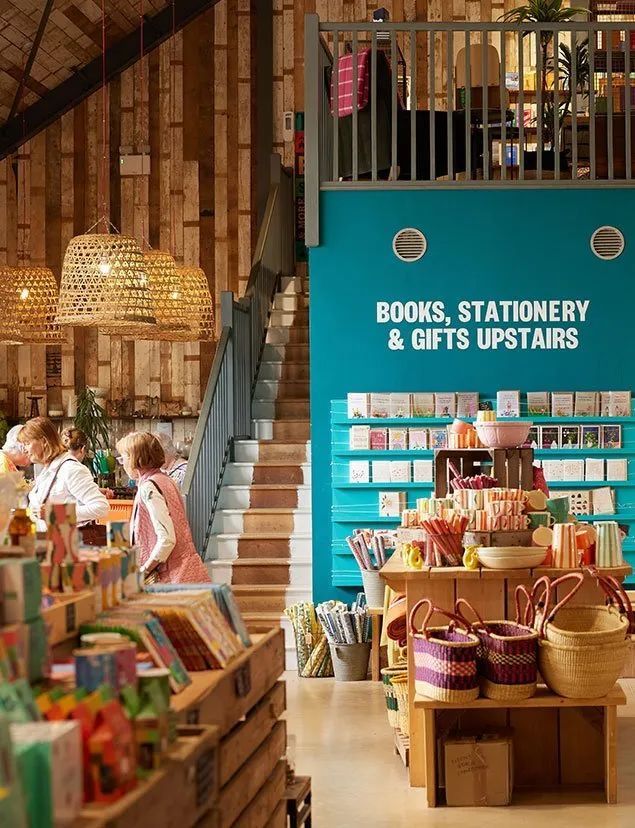 Shoppers browse inside the converted barn at Teals Farm Shop, where woven pendant lights hang over displays of cards, woven baskets, stationery and gifts, beside a teal wall with a sign reading “Books, stationery & gifts upstairs”.