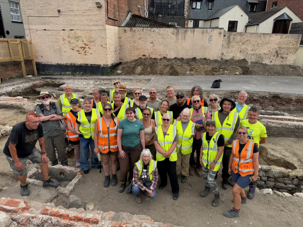 Dorset archaeology: a large group of smiling volunteers and archaeologists in hi-vis vests gathers among the excavated brick and stone foundations at the Weymouth Bowl site, during the dig led by Context One Heritage and Archaeology for Dorset Council.