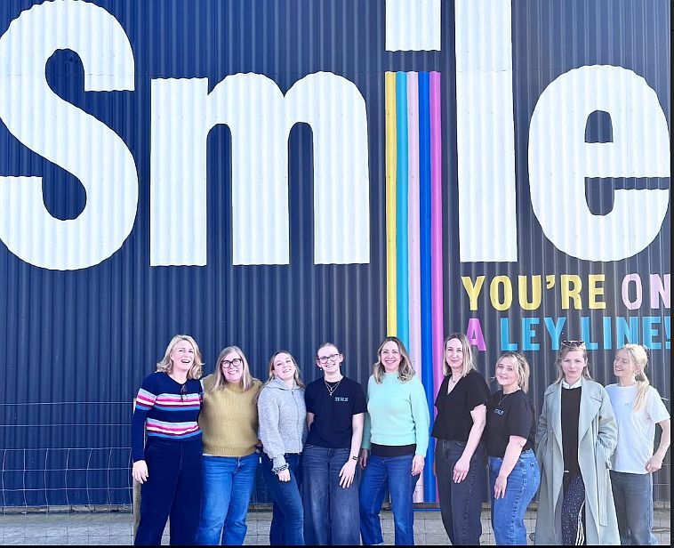 Nine smiling members of the Teals Farm Shop team stand in front of the barn’s giant “Smile, you’re on a Ley Line” sign, painted in white and rainbow lettering on dark blue corrugated metal.
