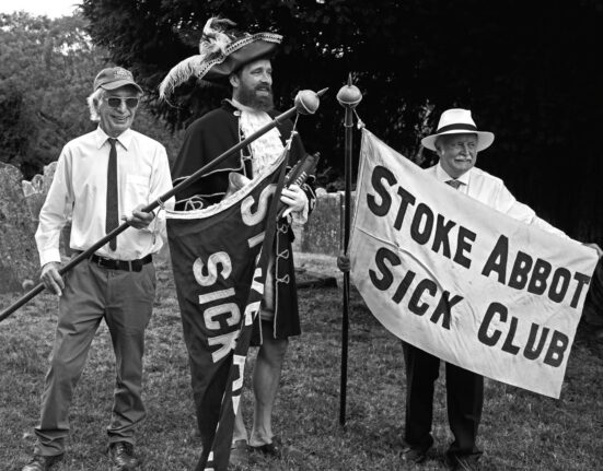 Three men hold Stoke Abbott Sick Club banners in a churchyard at the annual parade, one dressed as a town crier in a feathered tricorn hat.